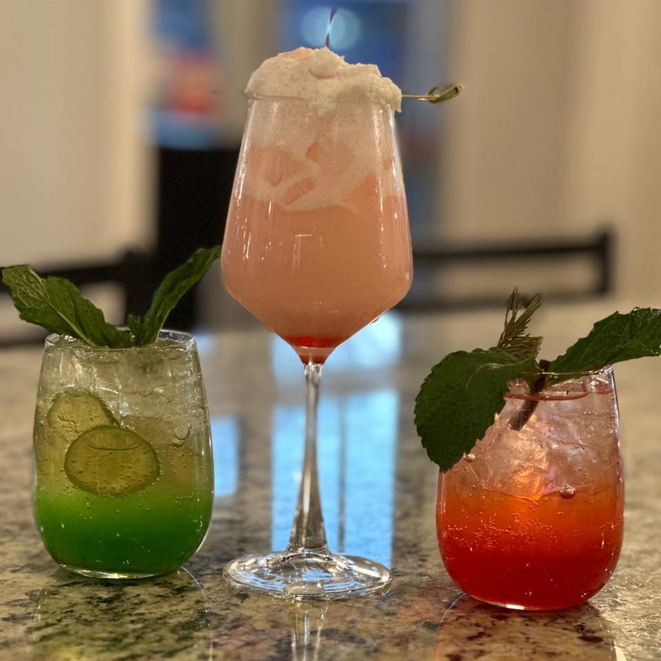Three colorful cocktails: green with lime, pink with foam, and red with mint, on a marble counter.