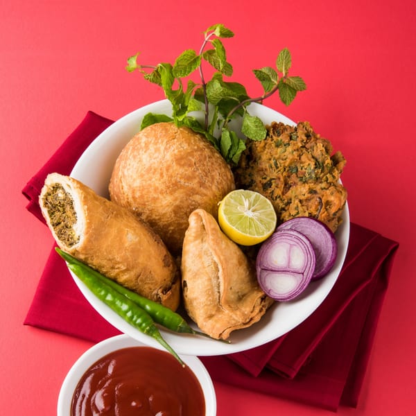 A white plate with Indian snacks like kachori, samosa, and pakora, garnished with mint, onion, and lime.