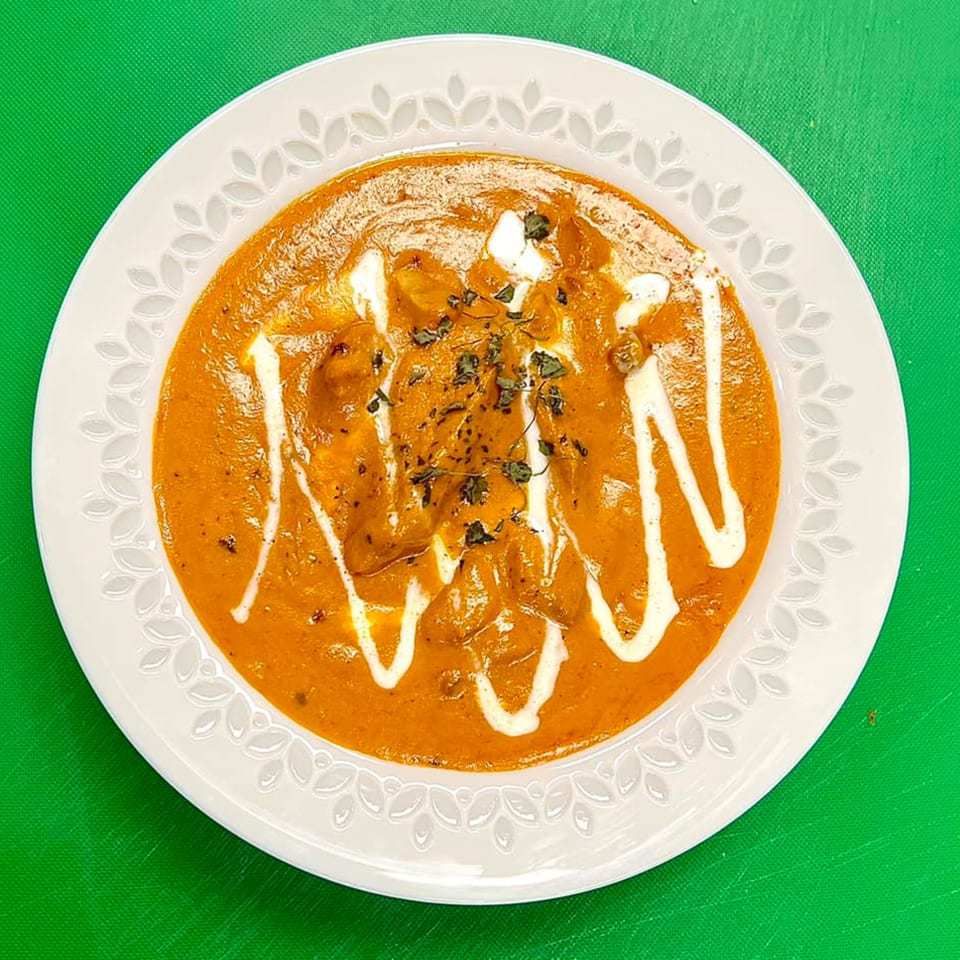 A white plate with a rich orange curry, drizzled with white cream and garnished with herbs.